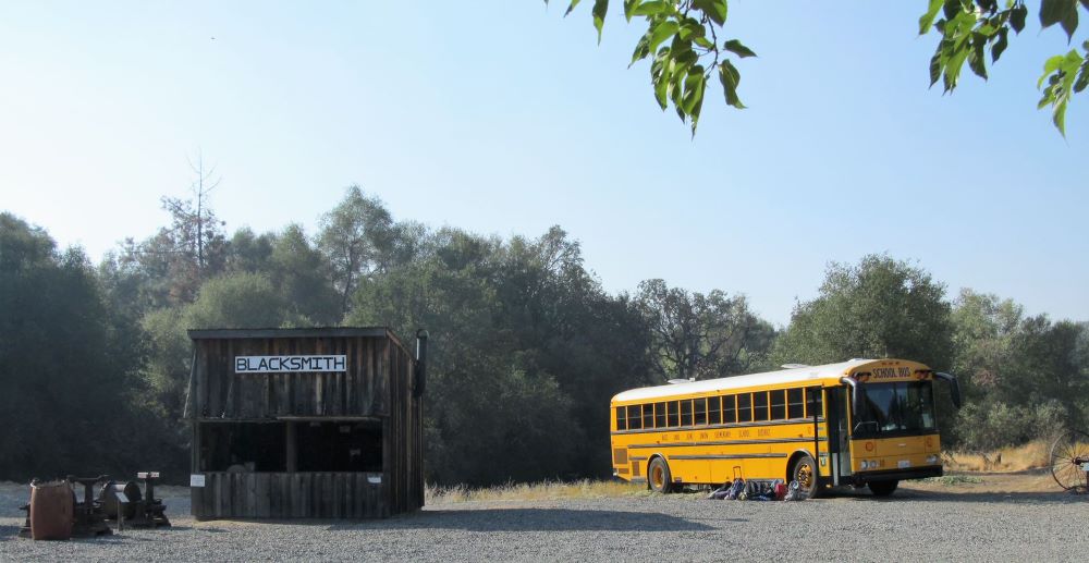 This is a photo of a school bus parked near the Blacksmith Shop. There is a pile of backpacks nearby. 